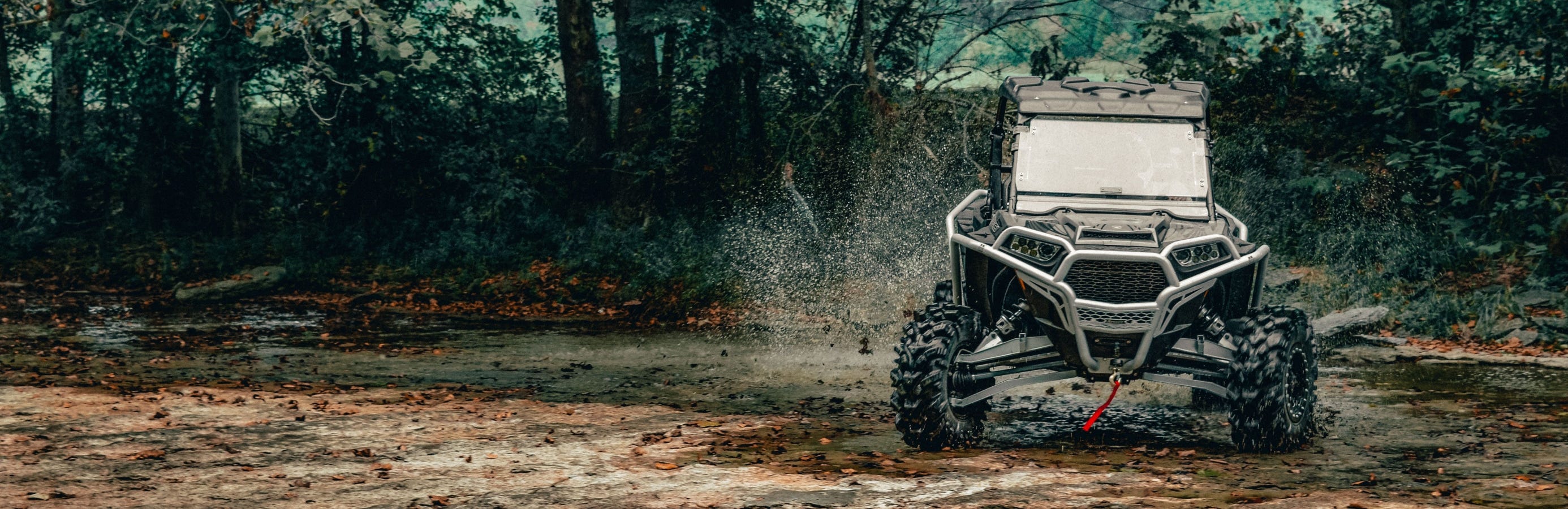 Black and silver Polaris RZR UTV tearing though the mud with a SuperATV flip windshield installed.